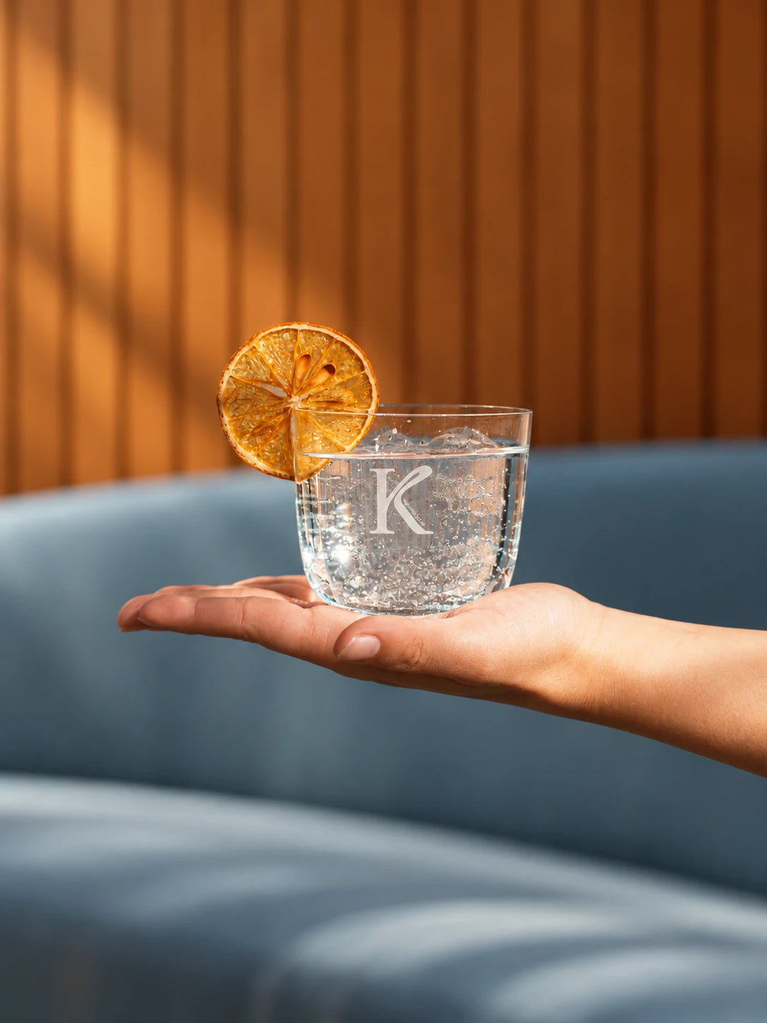 Hand hält ein Glas mit K-Monogramm und Orangenscheibe vor einem blauen Sofa